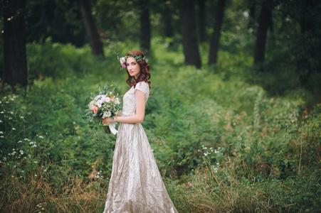 优雅的新娘头上戴着花环，穿着淡奶油的婚纱，站在夏天的森林里。