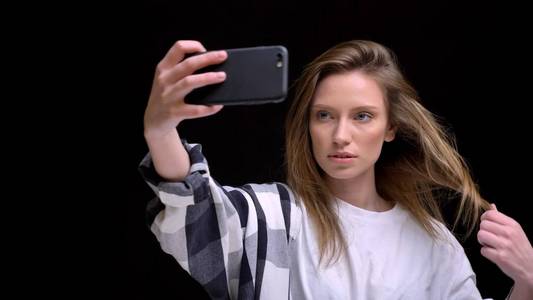 年轻的高加索长头发的女孩在衬衫制作自拍照片的肖像使用智能手机在黑色背景