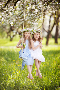 两个可爱的女孩在盛开的老苹果树花园的秋千上玩得很开心。 晴天。 儿童春季户外活动
