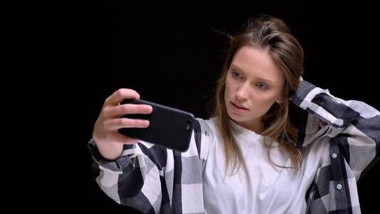 年轻的高加索长头发女孩的肖像格子衬衫触摸她的头发, 并在黑色背景上使用智能手机制作自拍照片