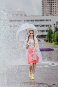 年轻漂亮的女孩与两个辫子在黄色的靴子和透明的雨伞站在喷泉附近