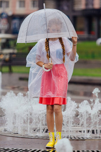 年轻漂亮的女孩与两个辫子在黄色的靴子和透明的雨伞站在喷泉附近