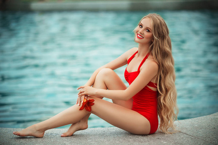 美丽的微笑的少女穿着红色泳衣与兰花在手在游泳池附近与蓝色的水休息。暑假