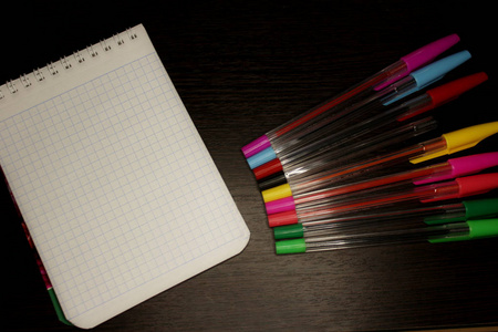 一本空白笔记本，上面有格子床单，在深色的木制背景上有一些多色笔。
