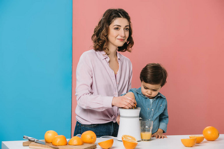 快乐的母亲和可爱的小儿子一起在双色背景下在榨汁机上榨鲜橙汁