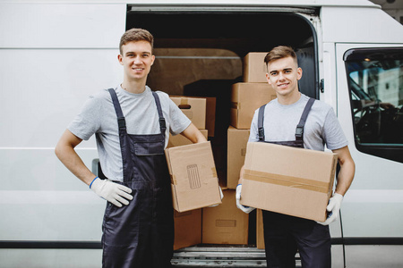 两个穿着制服的年轻帅气微笑的工人站在满是箱子的面包车旁边。房子移动, 搬运工服务