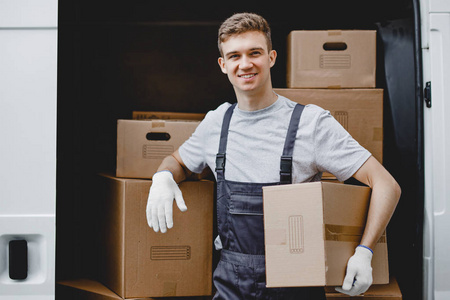 一名身穿制服面带微笑的年轻帅哥站在满车前, 手里拿着一个箱子。房子移动, 搬运工服务