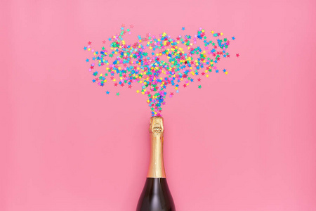 创意照片的香槟瓶与彩色纸屑在粉红色背景。 圣诞节周年庆新年庆祝概念平面图