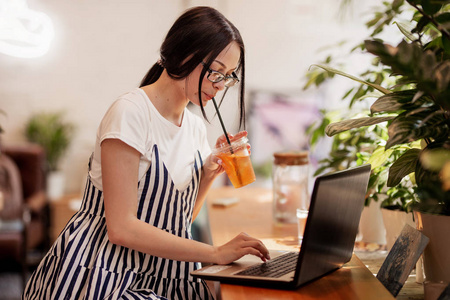 一个美丽的年轻苗条的女孩, 黑头发和眼镜, 穿着休闲的风格, 坐在桌旁, 在一个新的咖啡店与笔记本电脑