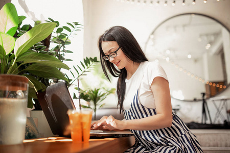 一个身材苗条的黑发女孩戴着眼镜, 穿着休闲的风格, 在舒适的咖啡店里在笔记本电脑上打字