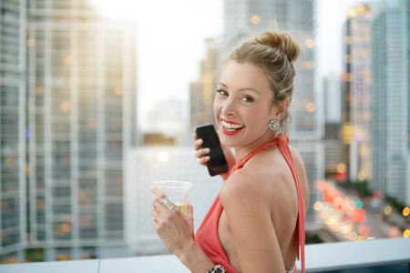吸引人的优雅年轻女子，在城市的屋顶酒吧里有饮料和手机