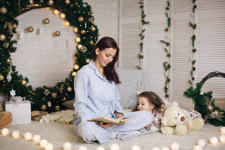 母亲给她可爱的小女儿在圣诞树附近看书