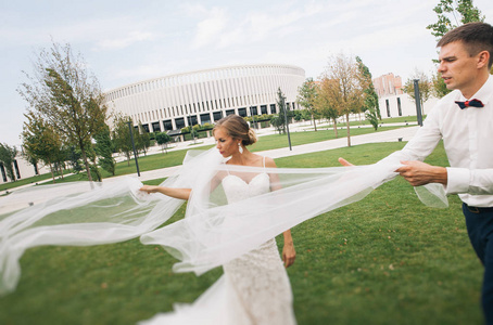 时尚的新娘和新郎在体育场附近的公园散步，戴上长长的面纱