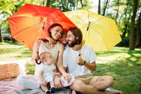 带着两个孩子的快乐父母在阳光下遮住他们的鲜红黄色雨伞下的草坪上休息