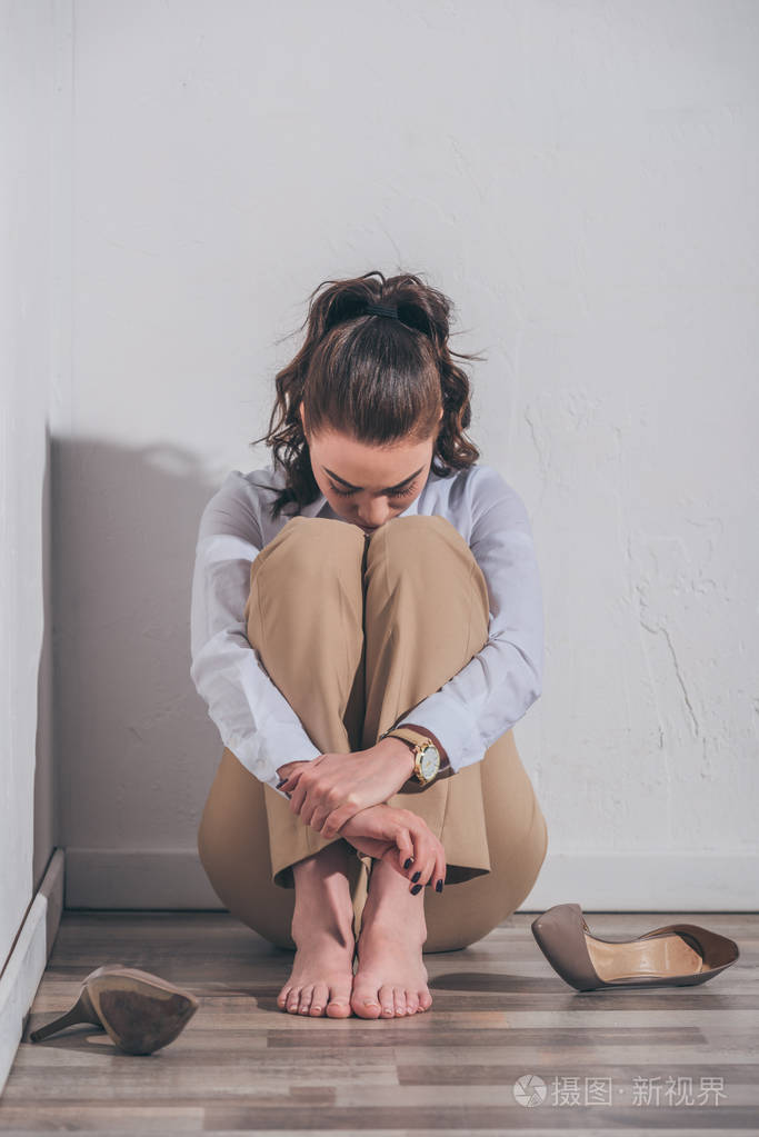 悲伤的女人穿着白色上衣和米色裤子,坐在地板上,抱着膝盖靠近墙壁