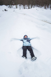 穿着裤子夹克手套和围巾的女孩躺在雪地里，在森林里堆雪天使