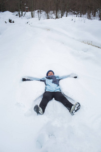 穿着裤子夹克手套和围巾的女孩躺在雪地里，在森林里堆雪天使