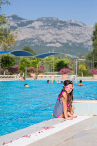 一个穿着泳衣的小女孩，穿着粉红色的泳镜坐在游泳池里，在山上的背景下