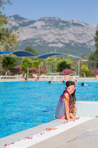 一个穿着泳衣的小女孩，穿着粉红色的泳镜，坐在土耳其山脉的背景上的游泳池上