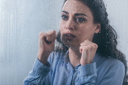 美丽的悲伤的女人，紧握的拳头透过窗户看着雨滴