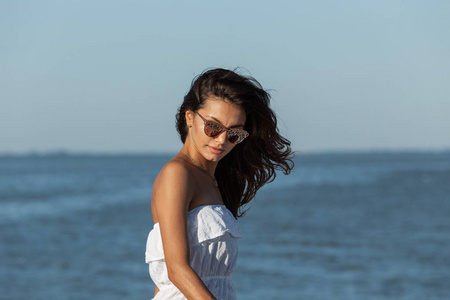 在阳光明媚的日子里, 美丽的黑发女孩的肖像, 穿着白色的衣服, 戴着太阳镜, 在海边