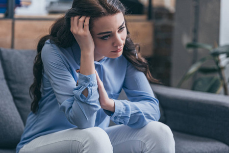 忧郁的女人，穿着蓝色衬衫，坐在灰色沙发上，望着远方的家，悲伤的无序概念