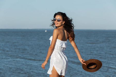 美丽的黑发女孩, 白色礼服和太阳镜拿着帽子在海边走近一个阳光明媚的日子