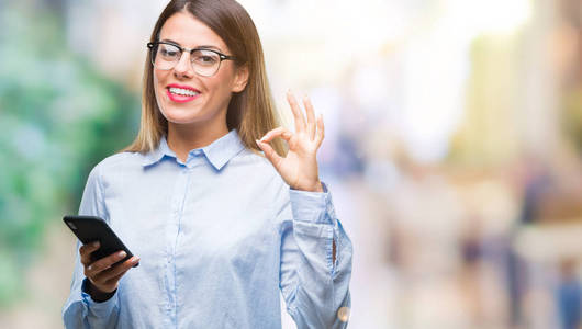 年轻漂亮的商务女性用智能手机在孤立的背景下发送短信，用手指做OK标志，优秀的符号