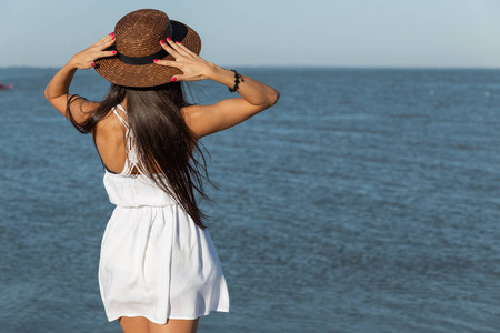 华丽的黑发女孩, 长头发, 白色礼服和棕色帽子站在海边的一个阳光明媚的日子