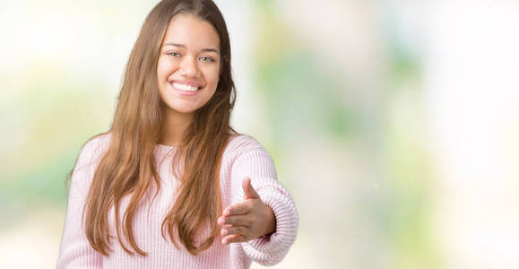 年轻漂亮的黑发女人穿着粉红色的冬季毛衣，在孤立的背景上微笑，友好地提供握手作为问候和欢迎。 成功的生意。