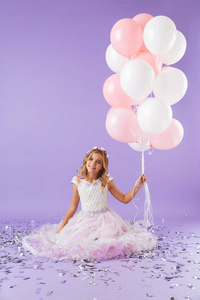 穿着公主裙的漂亮小女孩，独自坐在紫罗兰色背景下，手里拿着一束气球
