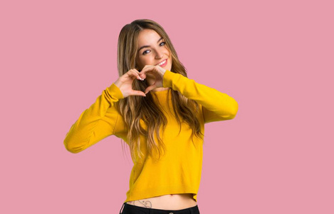 一个年轻的女孩，穿着黄色毛衣，用手做心的象征，用孤立的粉红色背景