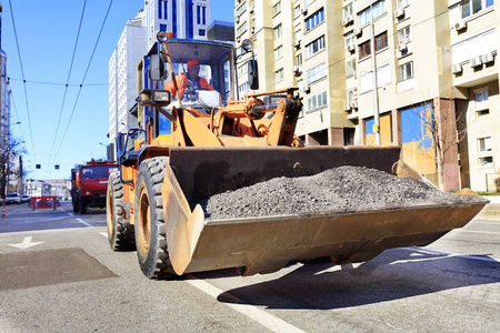 中午在一条城市街道上进行道路施工时，道路工人在重型建筑推土机铲斗中运送碎石