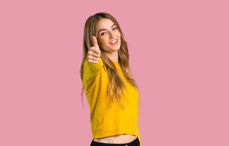 年轻的女孩，穿着黄色毛衣，竖起大拇指，因为在孤立的粉红色背景上发生了一些好事