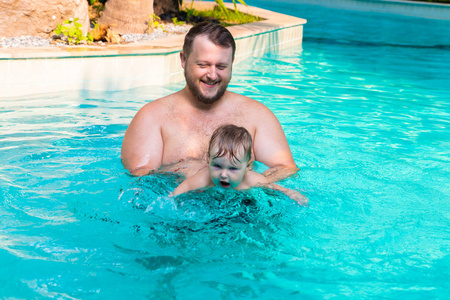 爸爸教一个孩子在游泳池里游泳。父亲和孩子在游泳池里玩得很开心。爸爸和女儿在棕榈树上的游泳池里游泳。父亲教女儿游泳。孩子们的概念