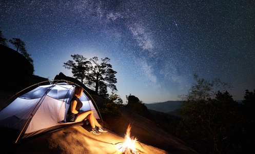 年轻女子徒步旅行者在篝火旁的岩石山顶上休息，夏季夜间还有发光的旅游帐篷