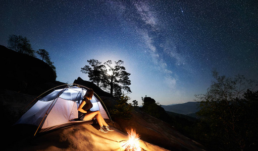 年轻女子徒步旅行者在篝火旁的岩石山顶上休息，夏季夜间还有发光的旅游帐篷