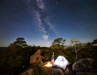 年轻女子徒步旅行者在篝火旁的岩石山顶上休息，夏季夜间还有发光的旅游帐篷