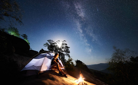 年轻女子徒步旅行者在篝火旁的岩石山顶上休息，夏季夜间还有发光的旅游帐篷