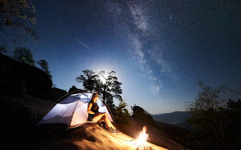 年轻女子徒步旅行者在篝火旁的岩石山顶上休息，夏季夜间还有发光的旅游帐篷