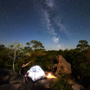 年轻女子徒步旅行者在篝火旁的岩石山顶上休息，夏季夜间还有发光的旅游帐篷