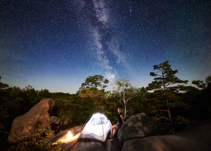 年轻女子徒步旅行者在篝火旁的岩石山顶上休息，夏季夜间还有发光的旅游帐篷