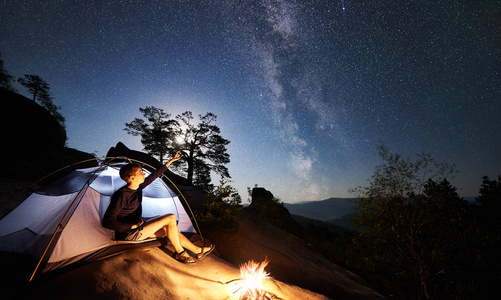 年轻的徒步旅行者在篝火旁的岩石山顶上休息，在夏季夜晚照亮旅游帐篷