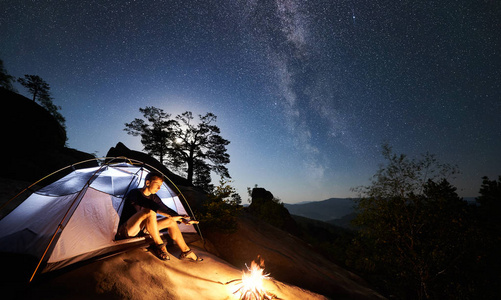 年轻的徒步旅行者在篝火旁的岩石山顶上休息，在夏季夜晚照亮旅游帐篷