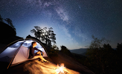 年轻的徒步旅行者在篝火旁的岩石山顶上休息，在夏季夜晚照亮旅游帐篷