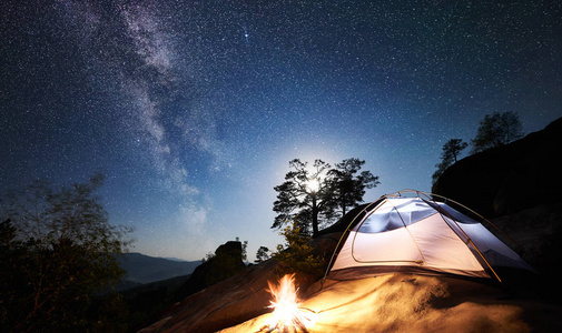 在充满星星和银河的神奇夜空下，白色旅游帐篷和篝火