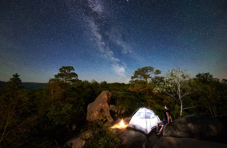 年轻女子徒步旅行者在篝火旁的岩石山顶上休息，夏季夜间还有发光的旅游帐篷