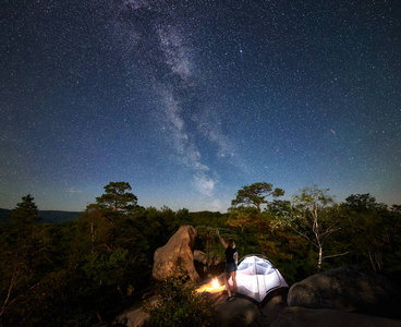 年轻女子徒步旅行者在篝火旁的岩石山顶上休息，夏季夜间还有发光的旅游帐篷