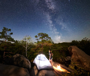 年轻女子徒步旅行者在篝火旁的岩石山顶上休息，夏季夜间还有发光的旅游帐篷
