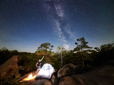 年轻女子徒步旅行者在篝火旁的岩石山顶上休息，夏季夜间还有发光的旅游帐篷